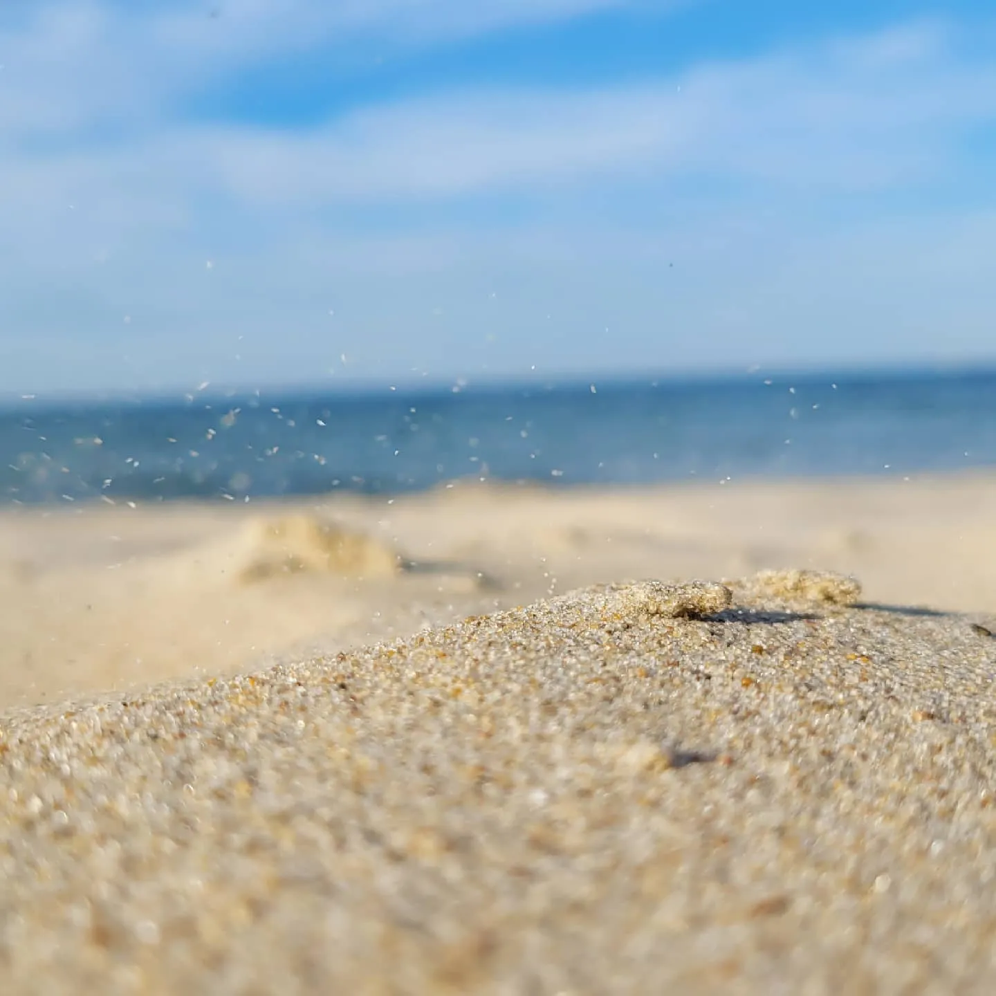 Macro of coarse beach sand in focus with grains kicked into the air, blurred sea and sky behind