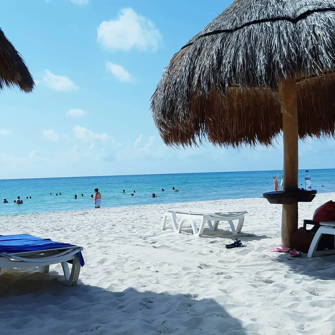 Thatched palapa umbrellas and sun loungers on white sand with swimmers in a calm turquoise sea