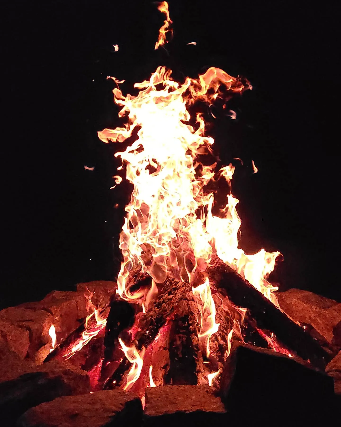 Campfire in a stone ring at night with tall flames and sparks against the dark