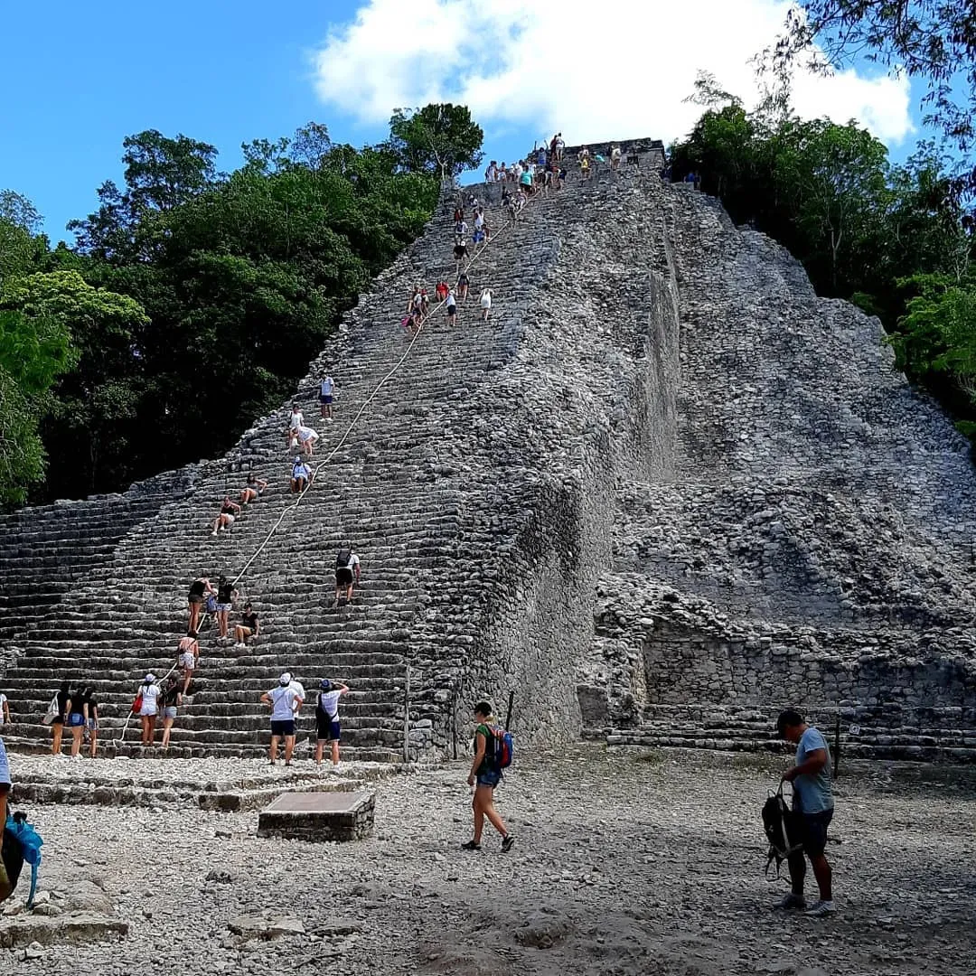 Visitors climbing the steep stone steps of the Nohoch Mul pyramid at Cobá, holding a rope line up the middle