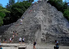 Visitors climbing the steep stone steps of the Nohoch Mul pyramid at Cobá, holding a rope line up the middle