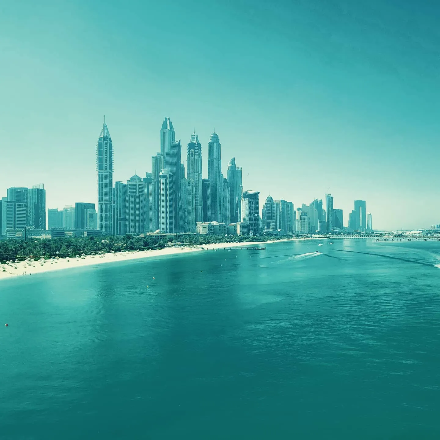 Dubai Marina skyscrapers lined along a beach and teal water, shot in a blue-tinted haze