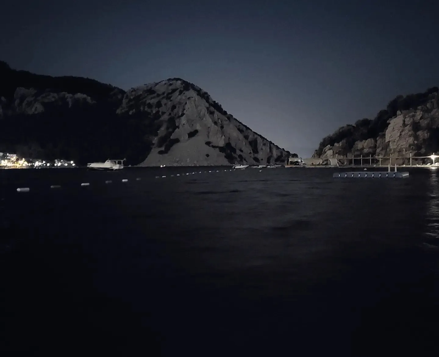 Dark bay after sunset with a rocky headland silhouette, buoy line, and village lights on the shore