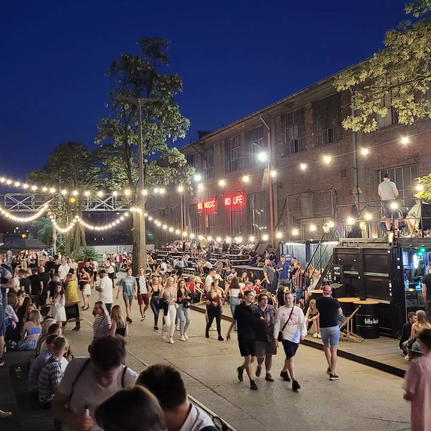 Crowded industrial courtyard at night with string lights, container bars, and a red neon sign reading "No music no life"