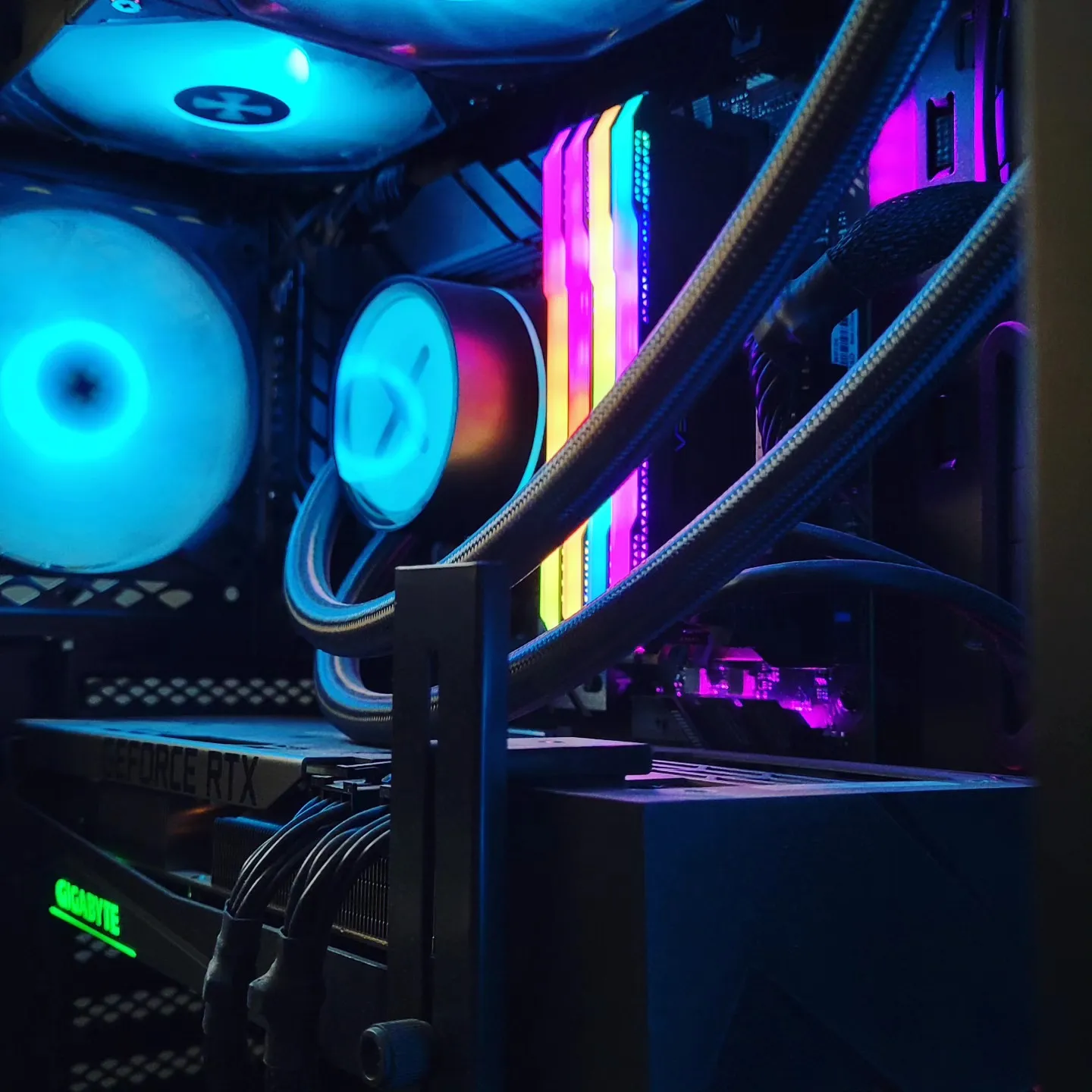 Inside of a PC case with cyan-lit fans, an AIO pump block, rainbow RGB memory sticks, and a GeForce RTX card