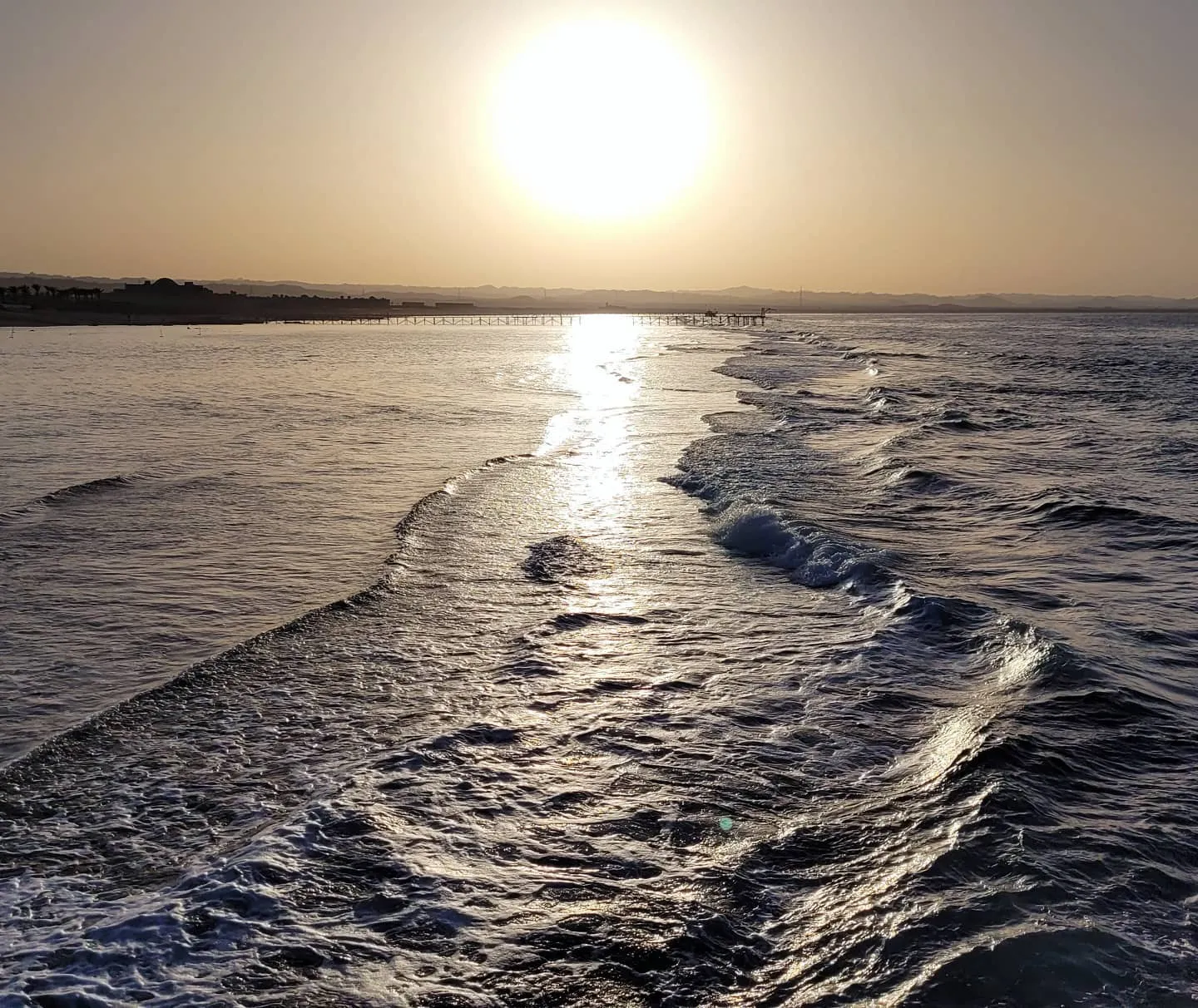 Low sun over the sea with waves rolling toward shore and a long pier silhouetted on the horizon