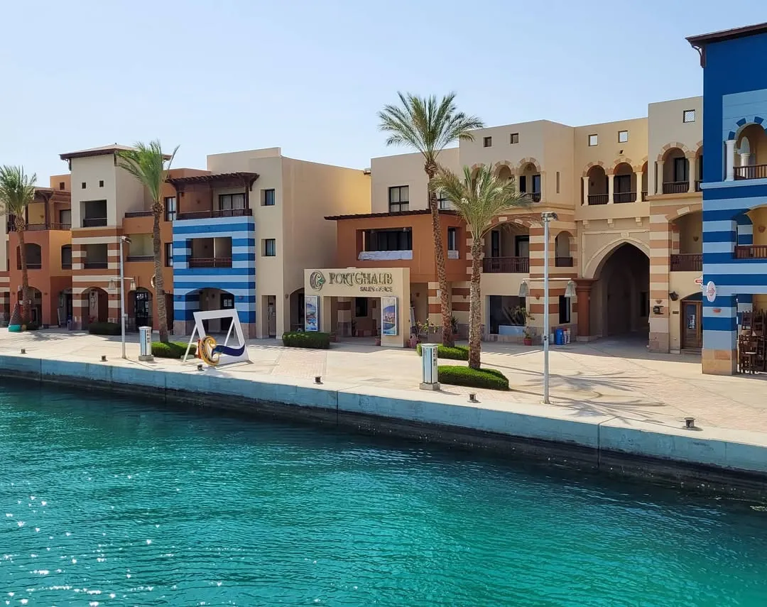 Waterfront promenade at Port Ghalib marina with striped orange-and-blue buildings and palm trees over turquoise water