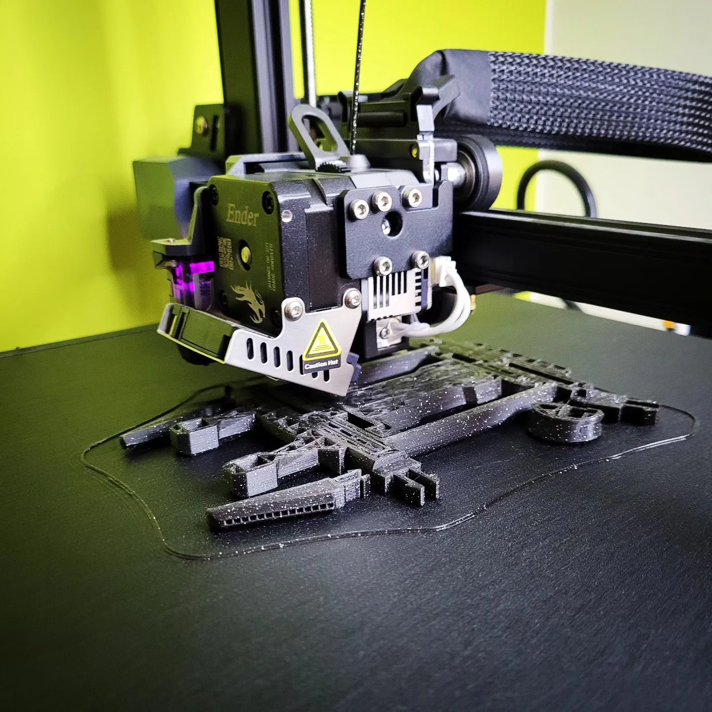 Ender 3D printer extruder mid-print over dark filament parts on a textured black bed, lime-green wall behind