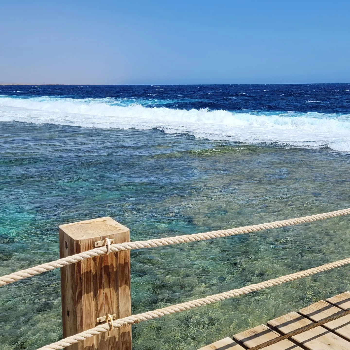 Wooden boardwalk with rope railing over a shallow reef, waves breaking on the deep blue sea beyond