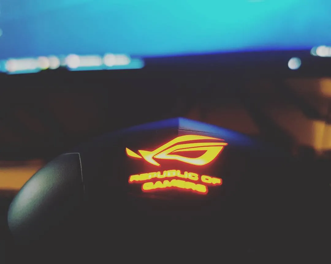 Republic of Gamers logo glowing red on a mouse in front of a blurred monitor at night