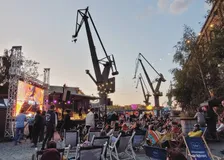 Open-air concert in deck chairs beneath the old Gdańsk shipyard cranes at dusk, stage lights on the left