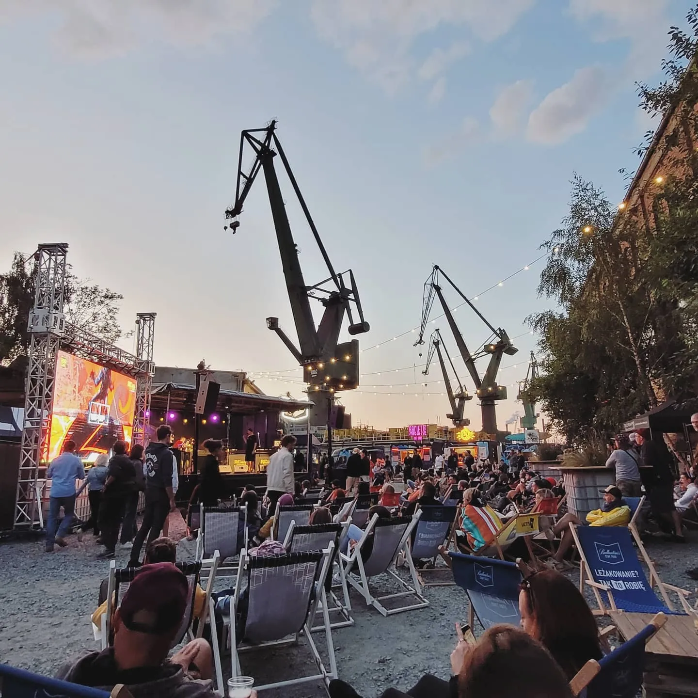 Open-air concert in deck chairs beneath the old Gdańsk shipyard cranes at dusk, stage lights on the left