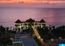 Thatched-roof restaurant on stilts over the sea at a pink-and-orange sunset, with a lit walkway leading to it
