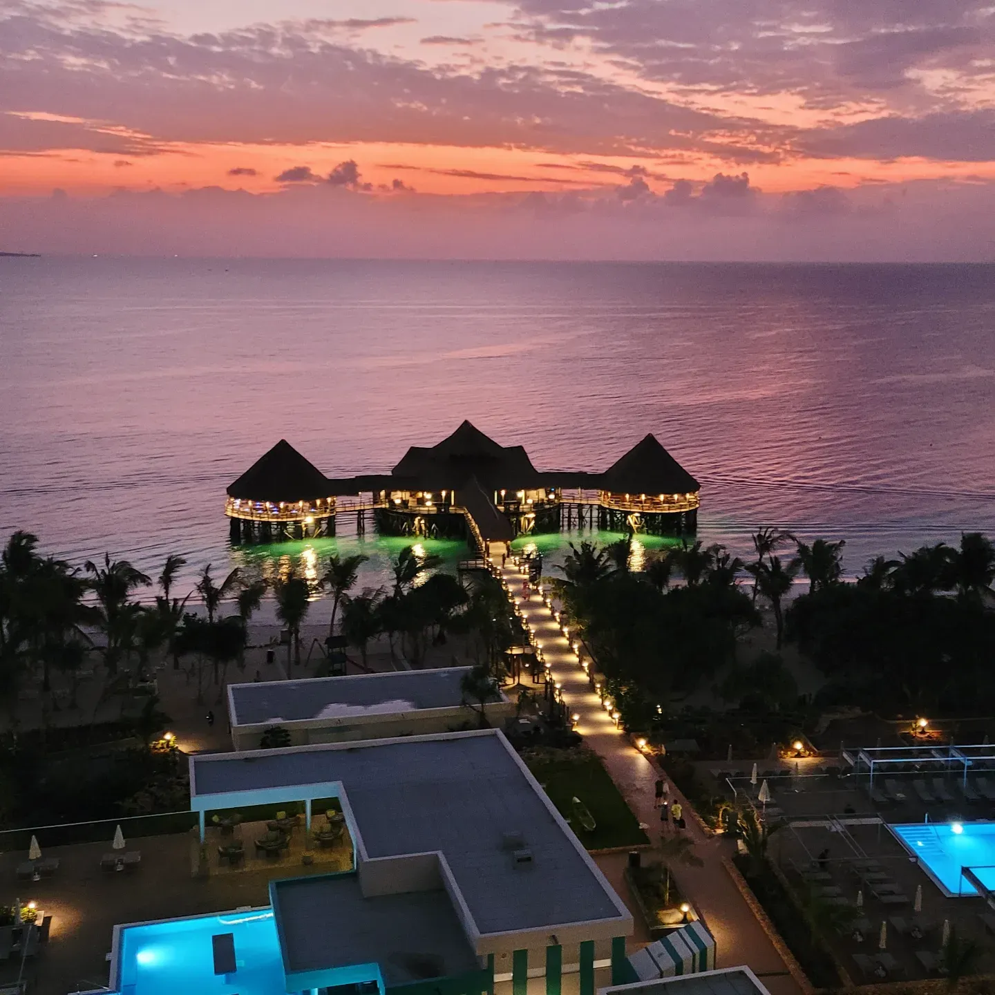 Thatched-roof restaurant on stilts over the sea at a pink-and-orange sunset, with a lit walkway leading to it
