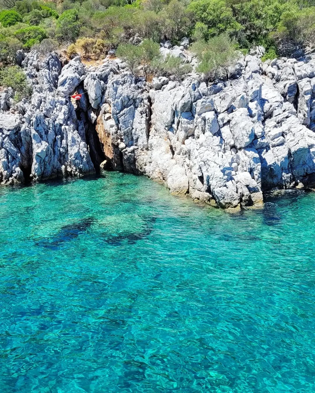 Small cove of glassy turquoise water below jagged white limestone rocks with a Turkish flag planted on top
