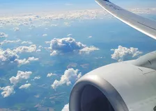 Aircraft wing and engine nacelle over scattered cumulus clouds and green patchwork fields far below