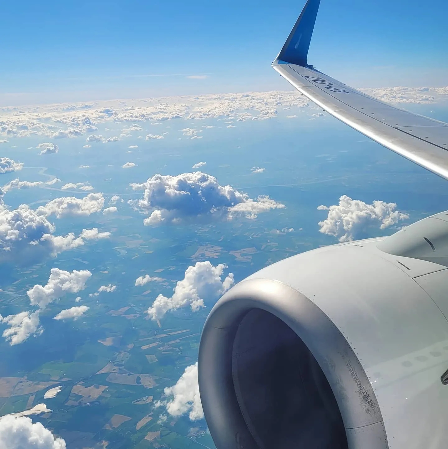 Aircraft wing and engine nacelle over scattered cumulus clouds and green patchwork fields far below